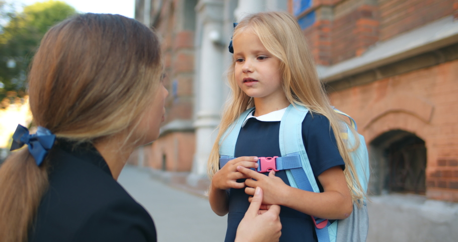 Close up of good looking young woman embracing her little kid near school building.Woman talking and stroking hair of her pretty daughter with bag and than waving at street | Shutterstock HD Video #1054432178