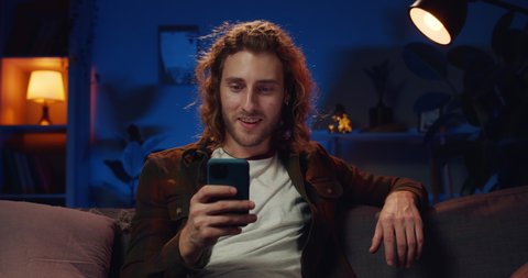 Millennial bearded man saying wow and making yes gesture while looking at smartphone screen. Good looking long haired guy winning while sitting on sofa. Concept of success and good news