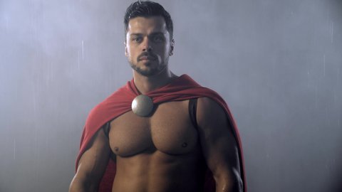 Front motion of spartan warrior standing under rain. Close up portrait of mature man in red cloak with water drops on muscular body posing in dark atmosphere. Ancient sparta, warrior concept.