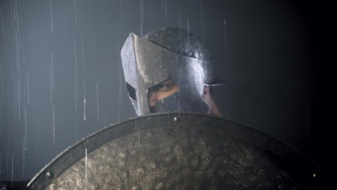 Close up motion of muscular spartan in armor and red cloak fighting with spear under rain. Man in helmet and historical costume holding shield and pointing weapon, looking at camera and smiling.
