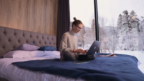 Portrait of attractive freelancer girl in glasses with a laptop on the bed in an eco-hotel with transparent walls behind which there is a winter forest. Travel and work concept with free schedule.