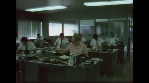 1960s: Man smokes and writes on notepad at desk in office space. Men and women work at desks in large office space.