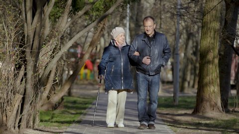 An elderly woman walks with her son enjoying an evening walk in park, talking and smiling. Man helps an elderly woman to walk. Couple of two generations - mother 81 years old and son 43 years old.