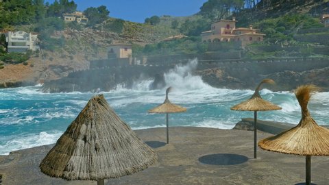 White topped waves crashing over the patio area, straw parasols and houses on hill in background.