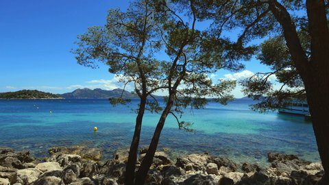 View of clear blue sea from a rocky coastline with trees in the foreground, mountains in the background.