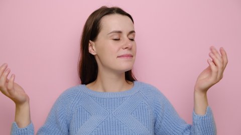 Cheerful young woman enjoying good smell or pleasant fragrance, feel calm breathe fresh air managing stress practicing exercises, wears blue sweater, peaceful girl isolated on pink studio background