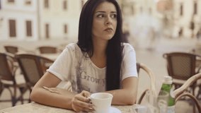 Young sad beauty woman is sitting with coffee at patio in HD VIDEO. She is waiting for message. In a moment message arrives and woman calms down and starts smiling. Walking people in background.