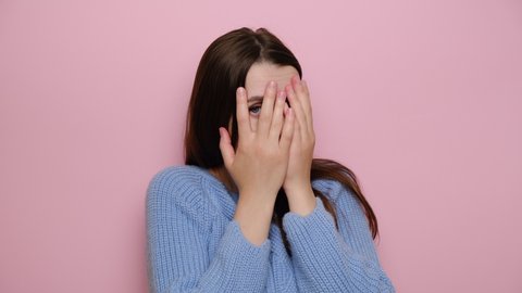 Scared young woman isolated on pink studio background hide behind hands, terrified brunette girl cover face peeping looking through fingers, curious female feel afraid peeking. People and phobia