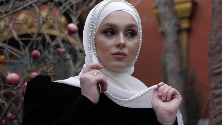 dreaming woman in stylish Arabian costume and white shayla decorated with pearls poses near pomegranate tree slow motion