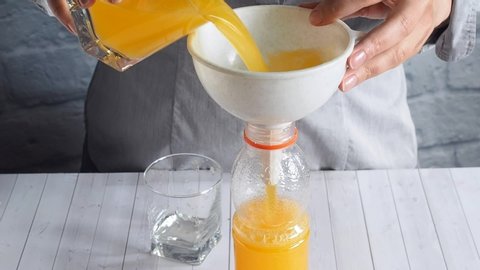 The girl at the table pours orange juice from glass glasses into a plastic bottle using a funnel.