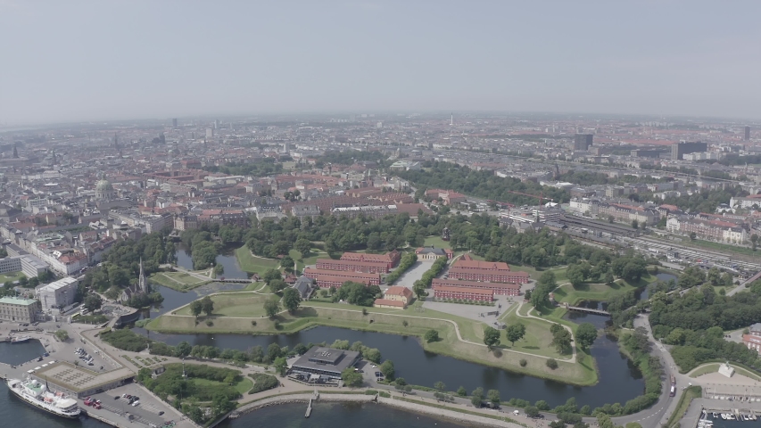 Copenhagen, Denmark. The star-shaped Kastellet fortress, one of the best-preserved pentagonal citadels in Northern Europe, dominates the frame with its five arrowhead bastions, earthen ramparts, and concentric water-filled moats. Built in the 1660s by Dutch military engineer Henrik Ruse, the fortress sits on the waterfront edge of central Copenhagen, its geometric precision visible from the sweeping aerial perspective. Long rows of red-roofed barracks line the central avenue within the walls, surrounded by manicured green parkland that doubles as a public space. The surrounding cityscape unfolds with dense urban blocks, a marina filled with docked boats, and the harbor waters of the Oresund Strait. The scene is captured under bright midday summer light with a clear pale sky, the flat northern daylight emphasizing the sharp angles of the bastions and the deep green contrast of the ramparts against the dark water. The camera executes a slow, steady orbit around the fortress, revealing the full pentagonal layout and its seamless integration into the modern city, Point of interest. D-Log.