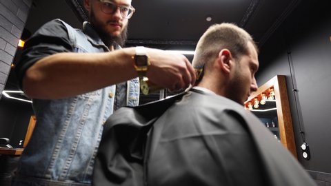 Trendy barber cuts bearded man's hair with a clipper in barbershop. Men's hairstyling and hair cutting in salon. Grooming the hair with trimmer. Hairdresser doing haircut in retro hair salon. Dolly