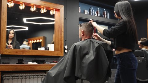 Attractive barber girl cutting man's hair with scissors and comb. Female hair cutter working serving client. Stylish man sitting in vintage barber shop chair. Hairstylist woman doing trendy haircut.
