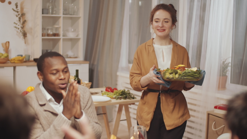 Young beautiful woman smiling and serving baked dish on table for cheerful multiethnic friends while having holiday dinner at home