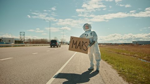 Man in Spacesuit is Standing at the Edge of a Road and Holding a Sign with Mars Written on it. Astronaut Looking to Hitchhike a Car. Spaceman in Futuristic Suit with Technological Panel on His Hand.