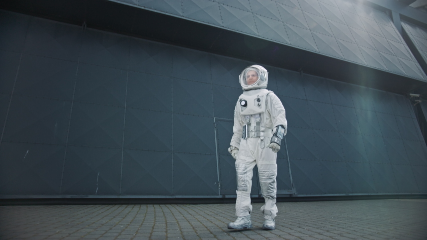 Low Angle Footage of a Confident Handsome Astronaut is Walking Towards the Camera from an Industrial Metal Wall. Man in Futuristic Suit with Technological Panel on His Hand.