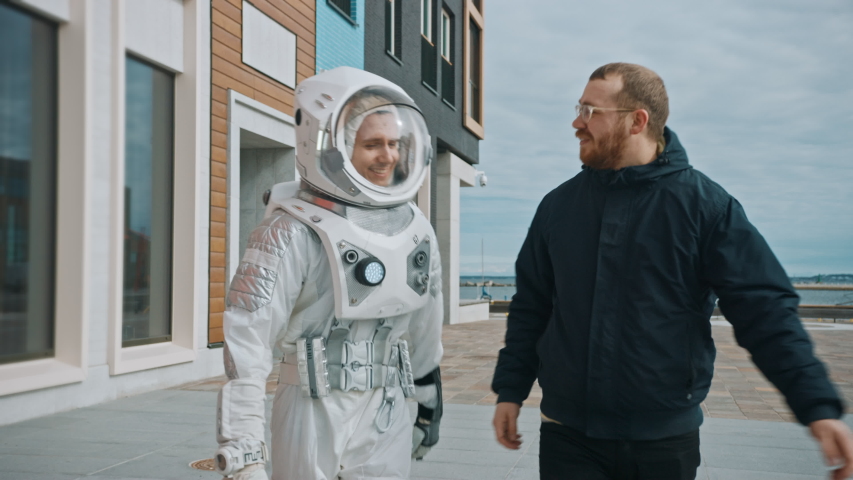 Ironic and Satirical Footage of Man in Spacesuit Walking on a Street with a Friend in Casual Clothes. Two Men Walk and Make a Pleasant Conversation. Man in Suit with Technological Panel on His Hand.