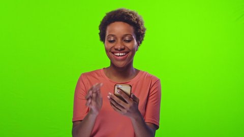 Positive African American woman with kinky short haircut in peach t-shirt surfs internet with phone at audition on green background