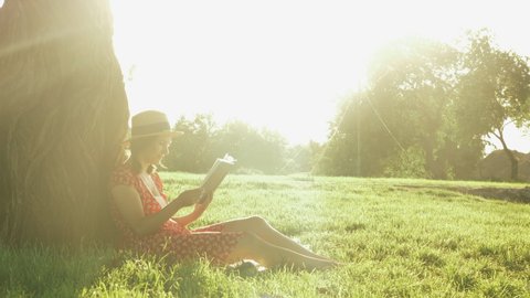 Cute young woman resting in park and reading book at sunny day in summer season. Girl leans on big tree while relaxing in park sitting on grass. Female student reading book diary in park