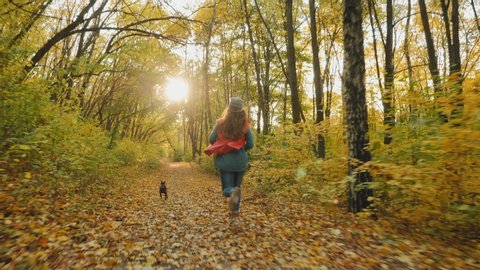 Rear view: Young girl running with her cute dog on the leash in the autumn park, slow-motion