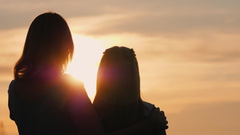 A woman gently hugs her daughter, watching the sunset together