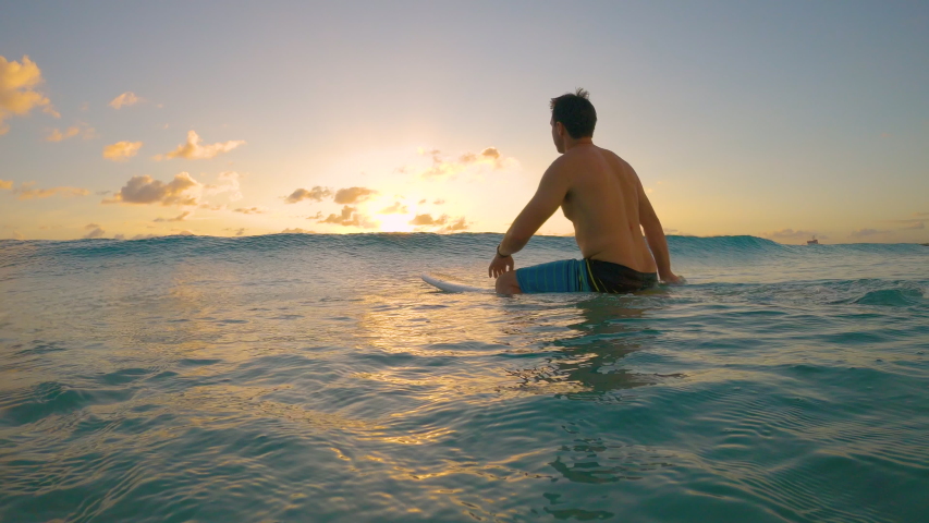 SLOW MOTION, LOW ANGLE, CLOSE UP: Caucasian man on surfing vacation in picturesque Barbados sits on his surfboard and watches the sunset. Male surfer is waiting in line up on an idyllic summer evening Royalty-Free Stock Footage #1054471697