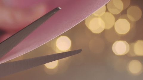 Close up of scissors cutting paper with defocused fairy lights in background. 4K resolution macro shot. Shallow depth of field. High quality audio recorded with condenser microphone.