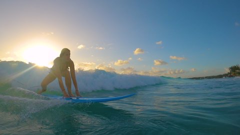 SLOW MOTION, LENS FLARE: Cinematic shot of a female beginner surfer riding a small wave to the tropical coast at sunset. Woman learning to surf stands up on her longboard and rides a wave at sunrise.