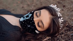 A video with a girl lying on the grass. She has a very beautiful mask on her face. She has a classic style crown on her head. The girl is dressed in black clothes.