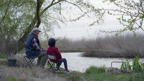 fishermen, an elderly man and his grandson in hats are sitting near shore and fishing small fish with a fishing rod in river background of trees and reeds on a warm day
