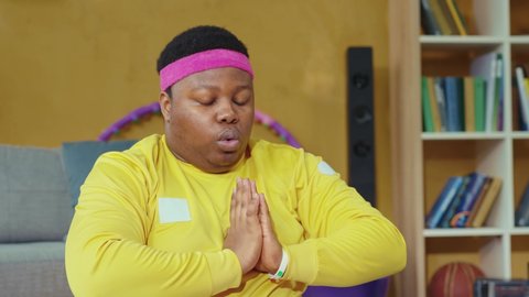 Calm african american guy practicing yoga asanas in the living room. Overweight relaxed young man sitting in lotus pose meditating at home.