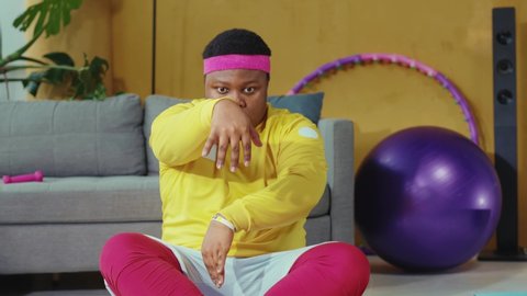 Funny-looking african young man wearing retro sportswear and doing yoga exercises meditation near sofa in living room. Home training. Sports, health, humor.