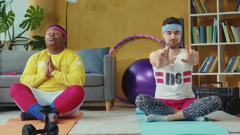 Comical male couple of funny friends jerks wearing retro style sportswear, sitting in lotus pose and practicing relaxing meditation.