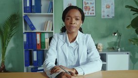 Attractive and friendly female doctor sitting in hospital office and talking by webcam with patient. Smart african physician explains treatment in medical videoblog. Online medicine.