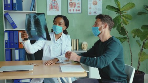 Female doctor in medical mask welcoming her patient, analyzing x-ray chest scan, feel sorry for negative test result. Unhappy patient. Medical examination. Bad diagnosis.
