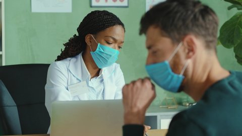 Afro-american doctor woman welcoming patient, covering face with medical masks, consulting in the office. Doctor appointment. Coronavirus. Pandemic.