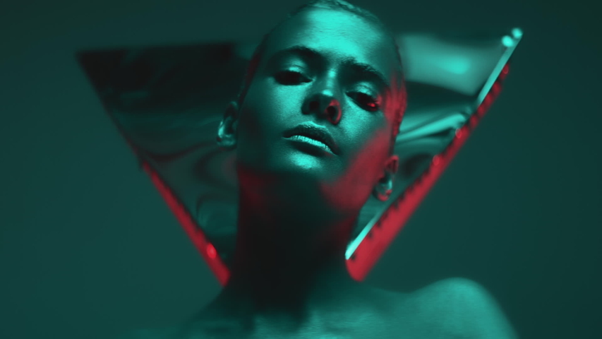 Gorgeous girl poses in studio, emotionally looking at camera. Portrait of beautiful young woman with shiny metallic makeup on face and body, illuminated by colorful neon light. Fashion model shooting