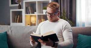 Smart young man with trendy haircut sitting on couch and reading interesting book. Bearded guy in glasses spending leisure time for self education.