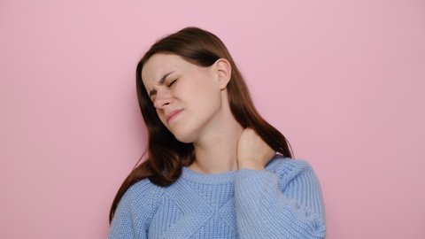 Tired upset young woman massaging hurt stiff neck, fatigued sad brunette girl rubbing tensed muscles to relieve joint shoulder pain, fibromyalgia concept, isolated on pink studio background