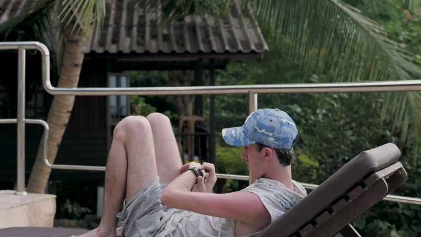 a young man with a smart watch on his hand sits under a palm tree on vacation. smart watch on the hands of a young man in a cap