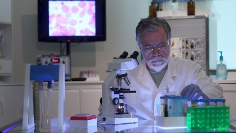 A cancer research scientist in a laboratory looking at a blood sample and taking notes with display of cancer cells on the monitor in the background.