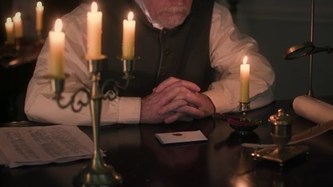 A scene from the 1800s era of a man sitting at a desk lit simply by candlelight who is opening a letter that has been sealed with a wax seal customary of the age.