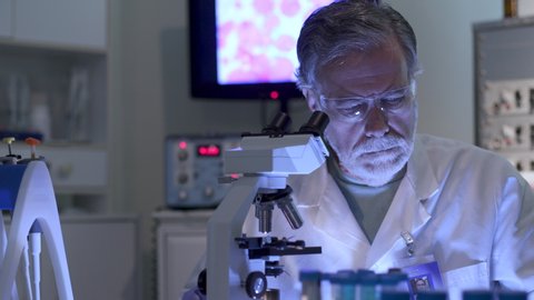 A cancer research scientist in a laboratory looking through a microscope with a display of cancer cells on the monitor in the background.
