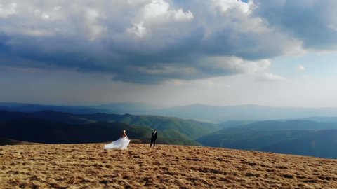 wedding couple bride and groom holding hands walking against the backdrop of amazing mountains and dramatic sky. Aerial view. Attractive blonde and brutal chat. In love and happy