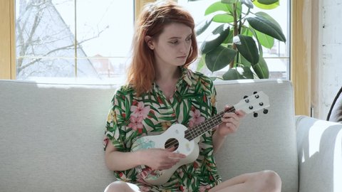 enjoying quarantine leisure, having fun, stay home concept: young pretty girl sitting on sofa plays toy guitar on sunny day. cute female in flowered shirt strumming ukulele on couch in bright room