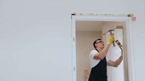 Male handyman carpenter at interior wood door installation with hammer and spirit level. Joiner assembling a doorpost or jamb between two rooms in new apartment