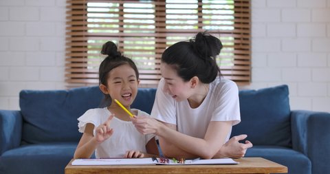 Young happy asian family mom or nanny helping cute kid daughter teaching toddler child learning drawing coloring picture with pencils together enjoy creative activity in living room at home.