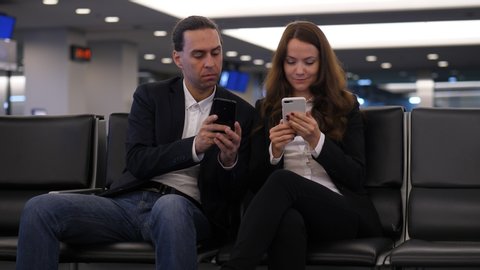Man and woman sit with phones at airport lounge, passengers waste time before flight. Guy show something on smartphone, woman shift closer and look to screen