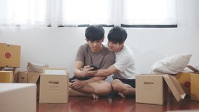 Asian couple man sitting, smiling and selfie via smartphone surrounding by carton package box storage for move in empty new home. LGBTQI relationship pride month and relocating a new house concept