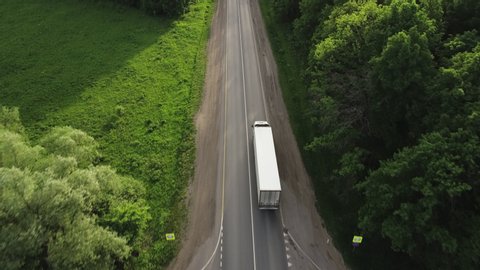 One Semi Truck with white trailer and cab driving / traveling alone on dense flat forest asphalt straight road, highway top view follow vehicle aerial footage / Freeway trucks traffic
